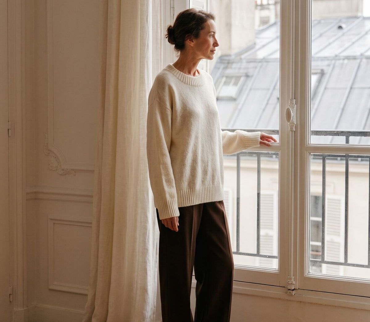 Femme en pull en maille écru ouvrant une fenêtre avec vue sur les toits parisiens
