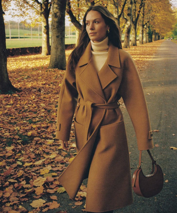 Femme en manteau camel ceinturé dans une allée bordée d'arbres en automne
